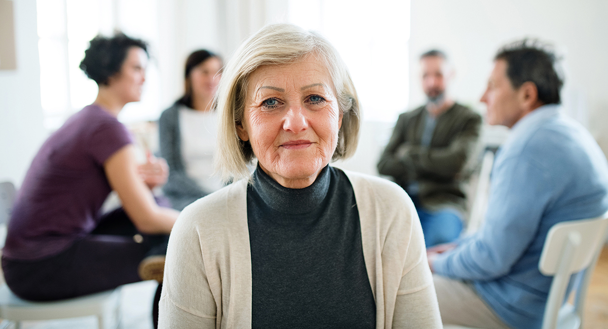 Photo of an older woman facing the camera with a support group gathered in the background.