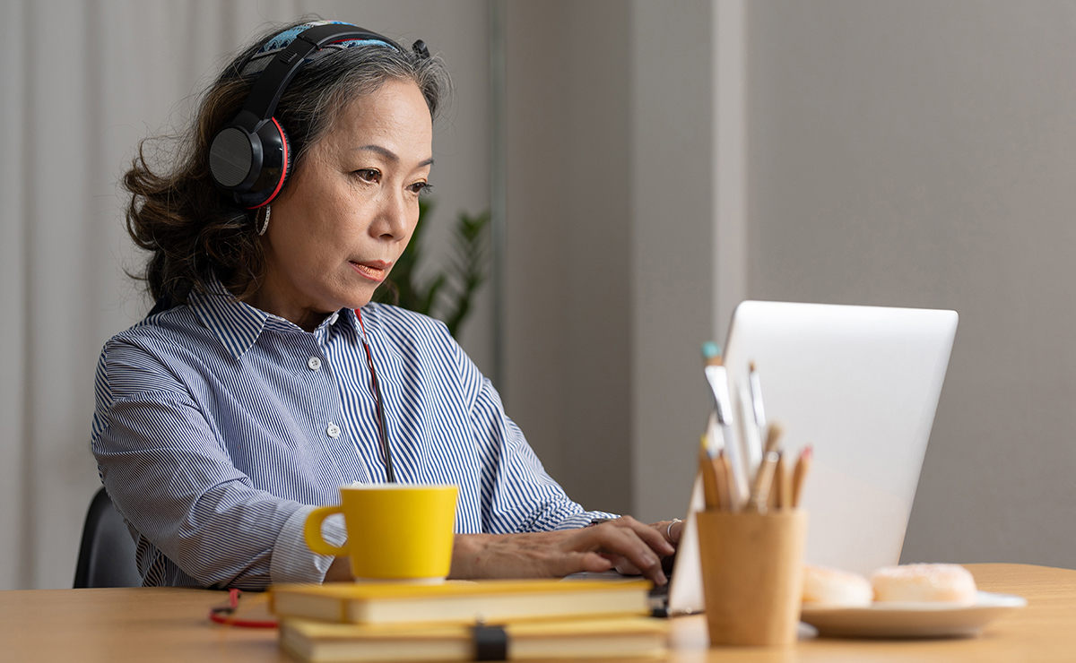 Photo of a woman seated at a desk wearing headphones while watching something on her laptop.