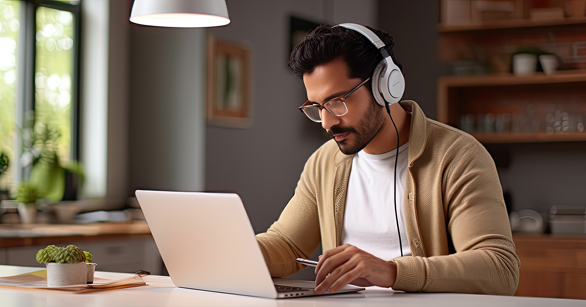 Photo of a man wearing headphones and staring intently at his laptop while seated in his kitchen.
