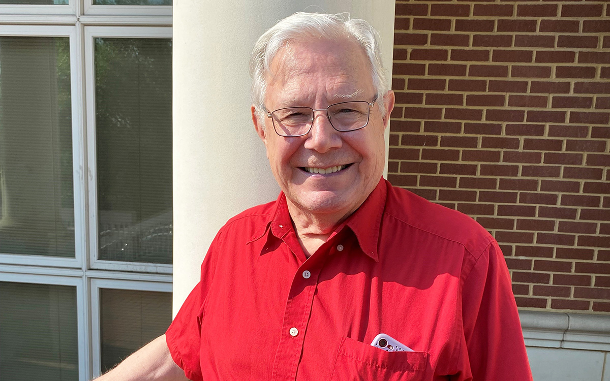 Alan Cole smiling and wearing a red shirt while standing in front of a white column outdoors.