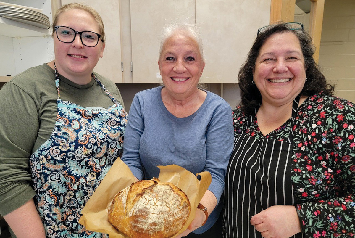 Three women stand smiling with the center woman holding a rustic loaf of bread. 