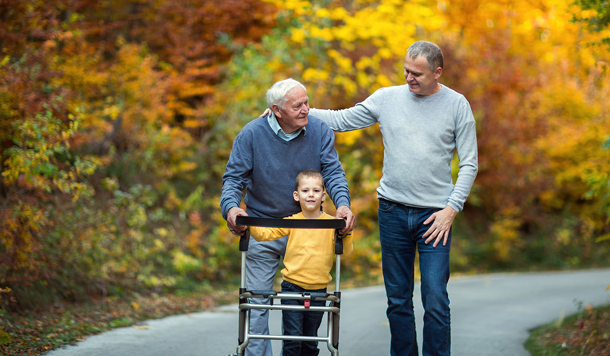 An older man with a walker walks down a road with a younger man and a boy. In the background is brilliant fall foliage.