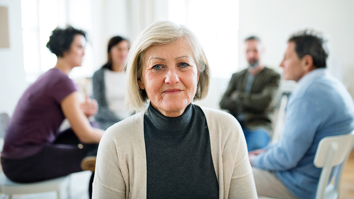 An older white woman looks directly at the camera while a group of people gather in a circle in the background.