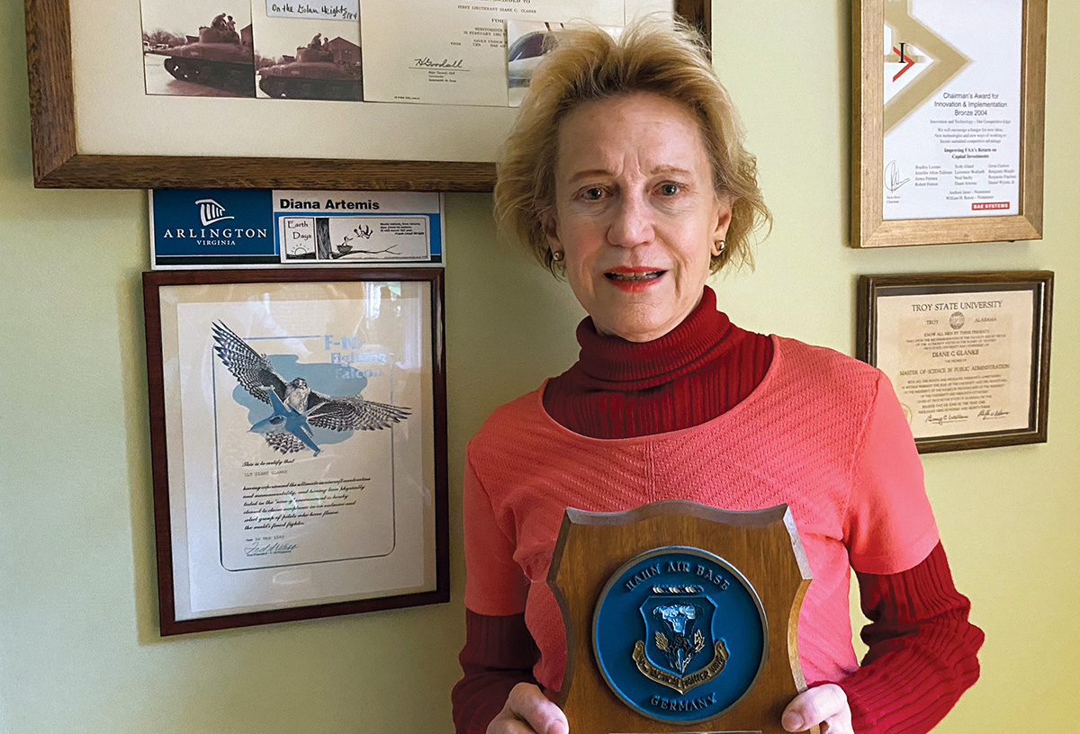 Diana Artemis in her home, standing in front of a wall of framed photos and certificates.