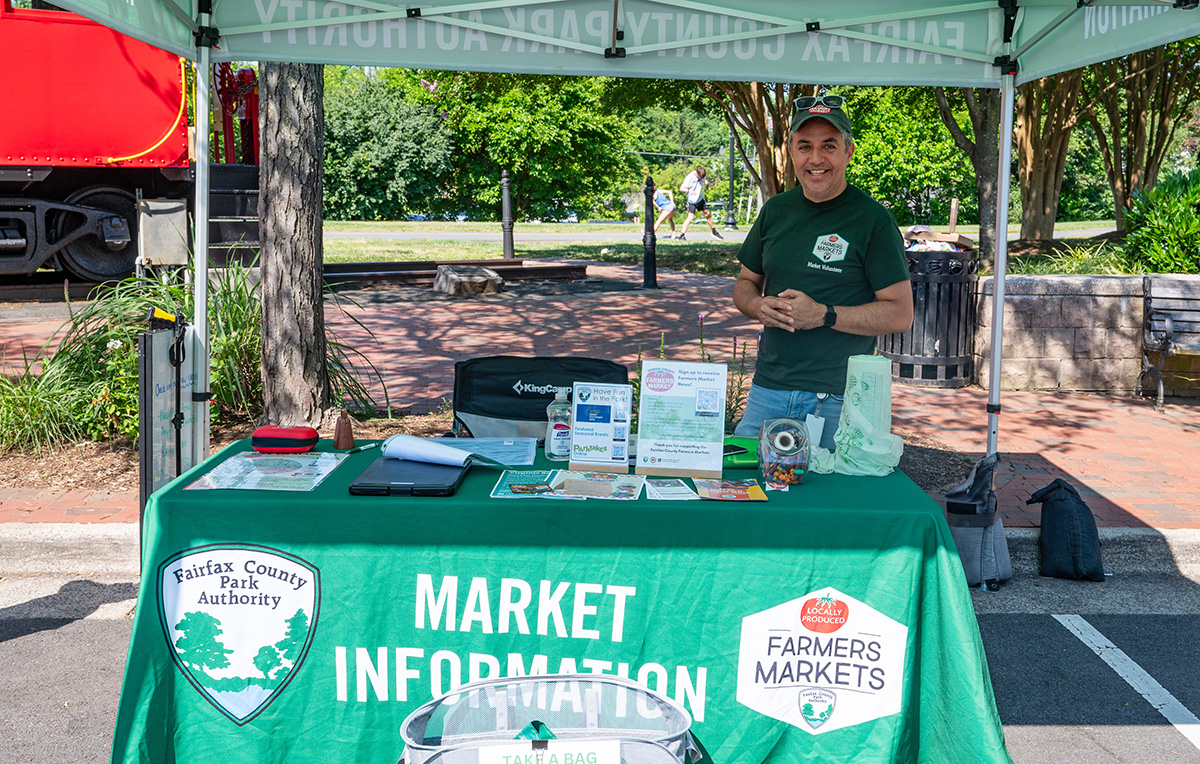 A man volunteers at a Farmer's Market table situated under a canopy.
