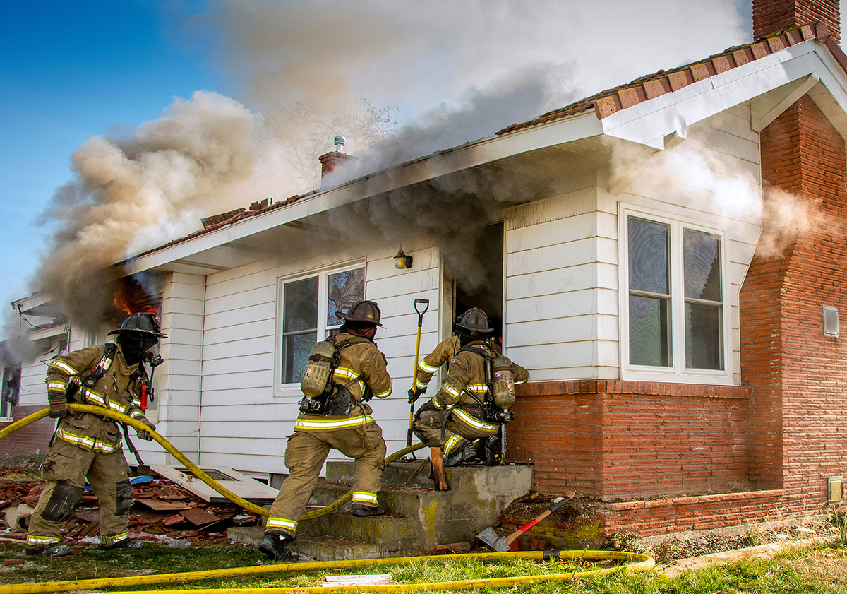 Firefighters entering a home with smoke billowing from the windows.
