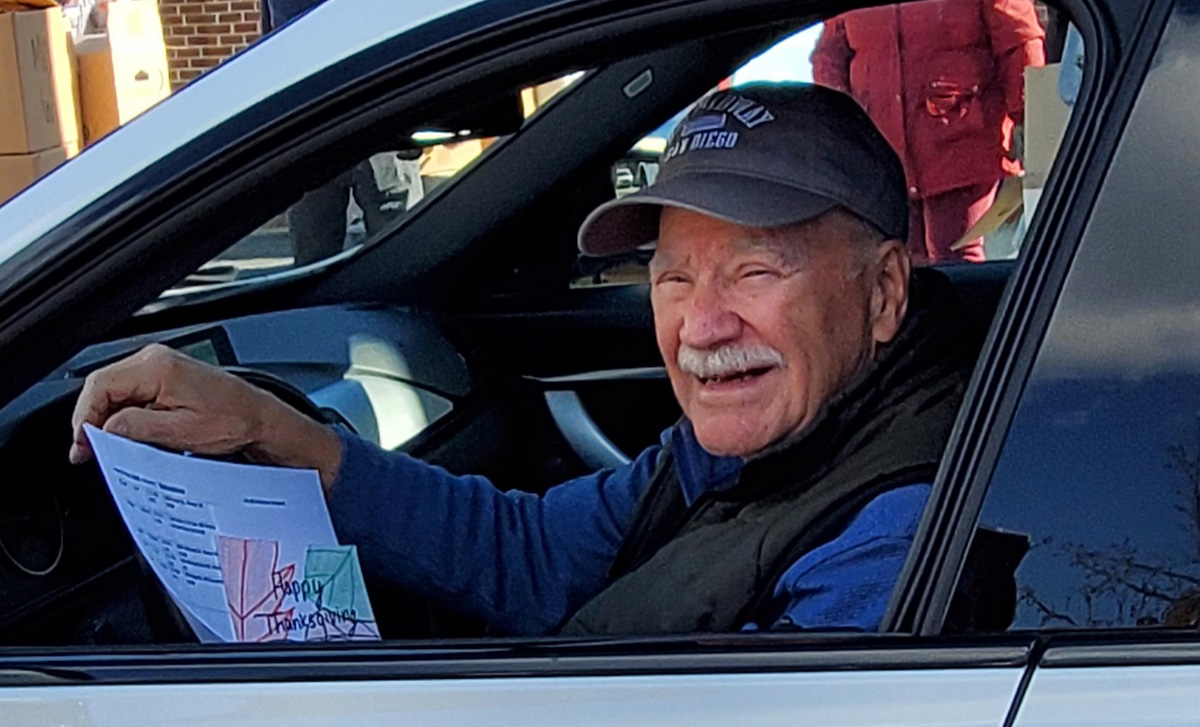 Volunteer George Bishop sits in the driver's seat of a car wearing a baseball cap and smiling.