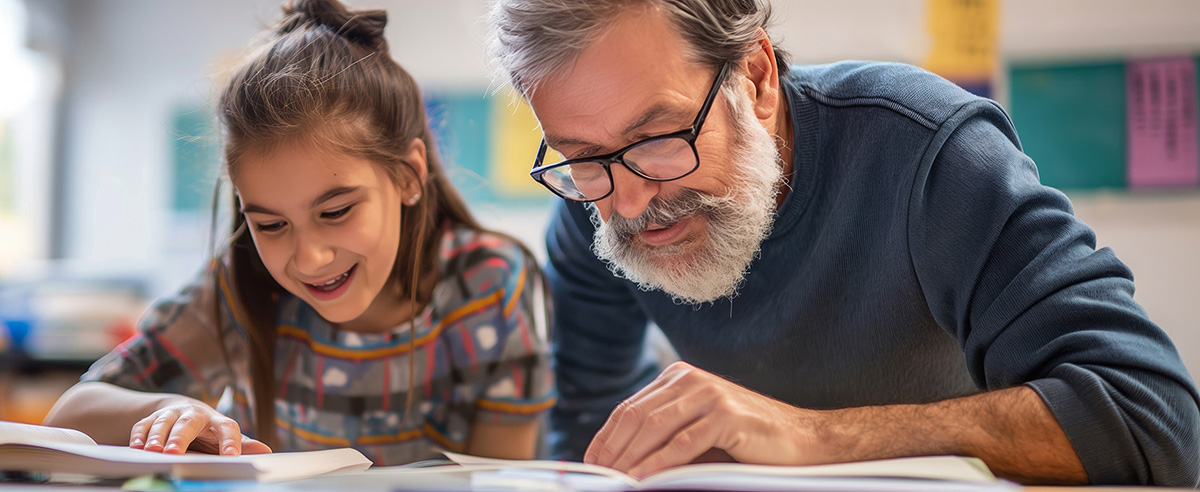 An older man and a girl read books together while seated at a table.