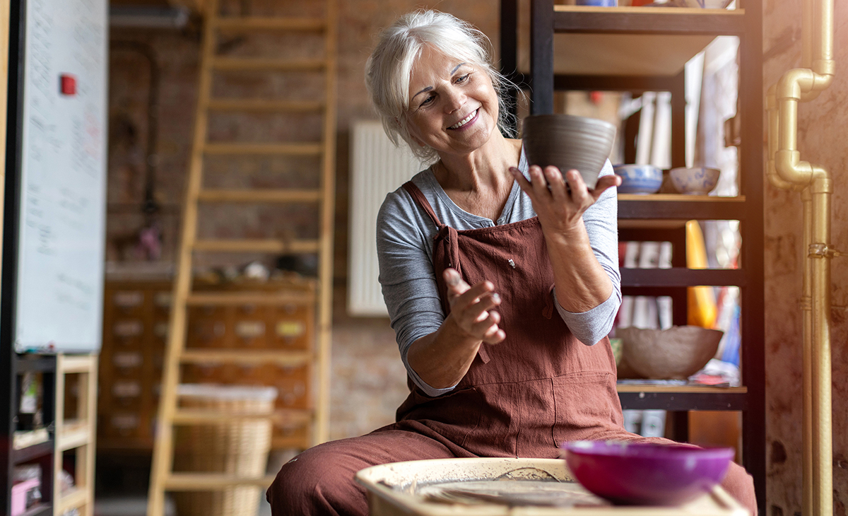 An older white woman seated at a pottery wheel in an art studio holding a newly made pot.
