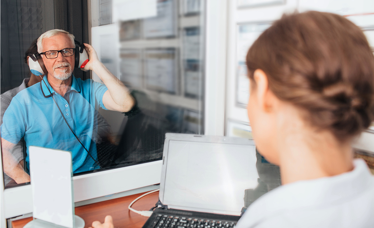 Older white man sitting in a sound booth undergoing a hearing test while a young woman technician sits outside of the booth.