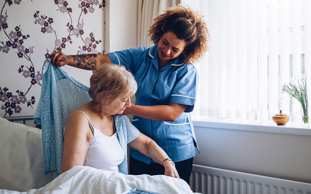 A health aid wearing blue scrubs helps an older woman dress while she is seated on a bed.