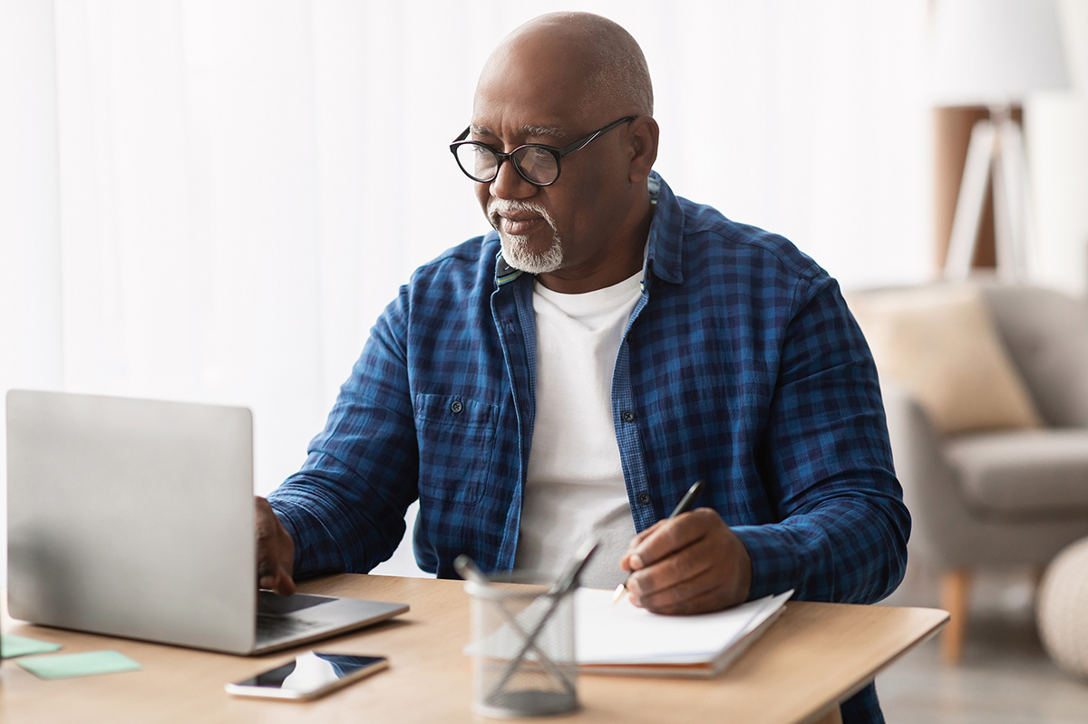 Photo of an older African American man seated at a desk with a laptop open in front of him. His hand holds a pen and is poised to write over a notebook.
