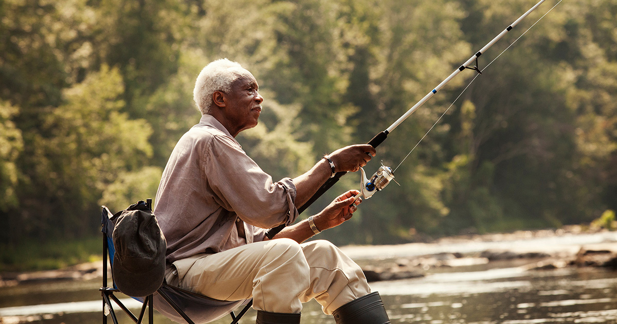 An older African American man sits in a lawn chair holding a fishing pole next to a river.