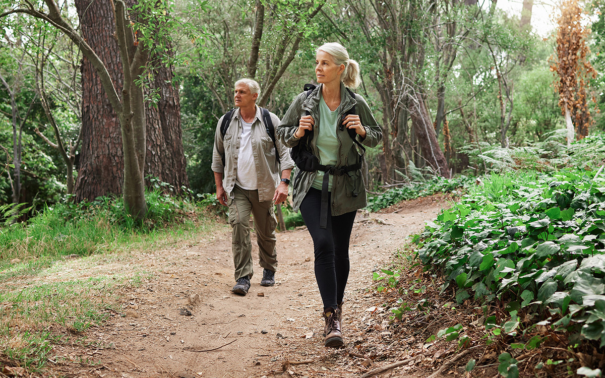 An older couple wearing backpacks hikes in a wooded area.