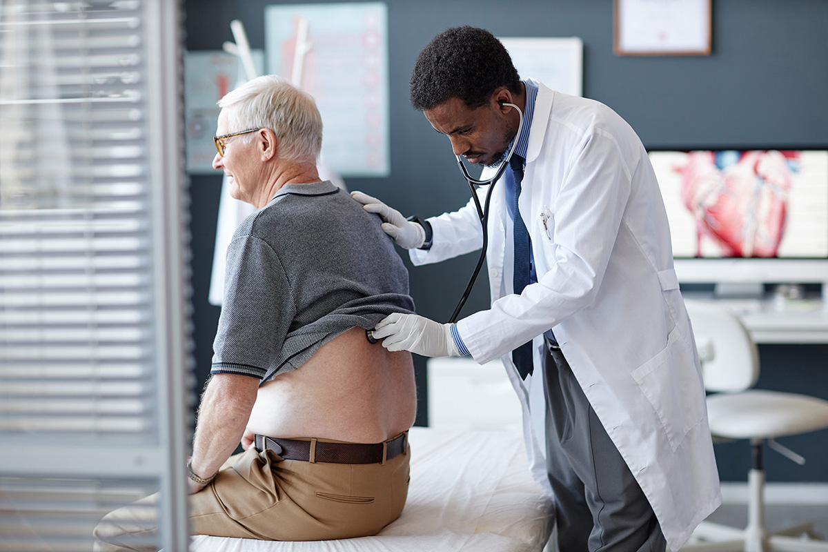 An older man sits on an exam table while a doctor listens to his lungs with a stethoscope.