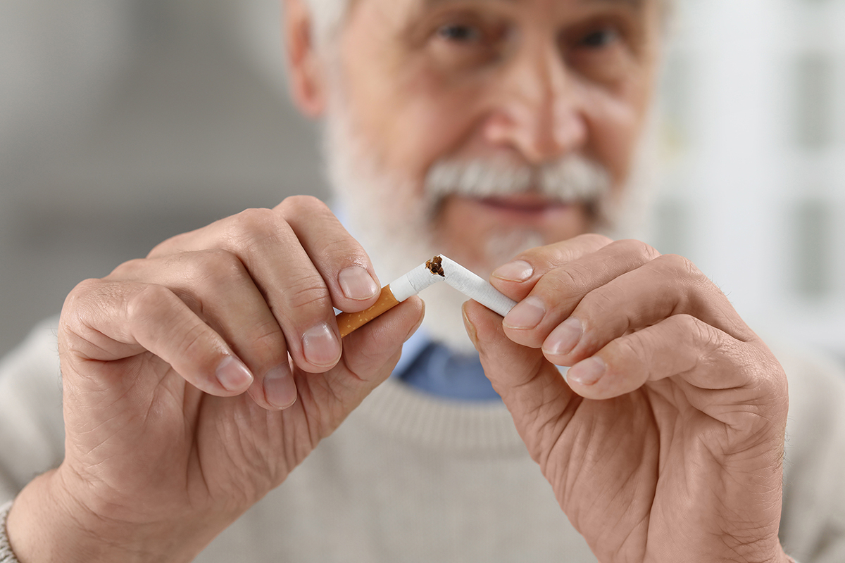 Photo of an older bearded man wearing a white sweater and breaking a cigarette in two pieces.