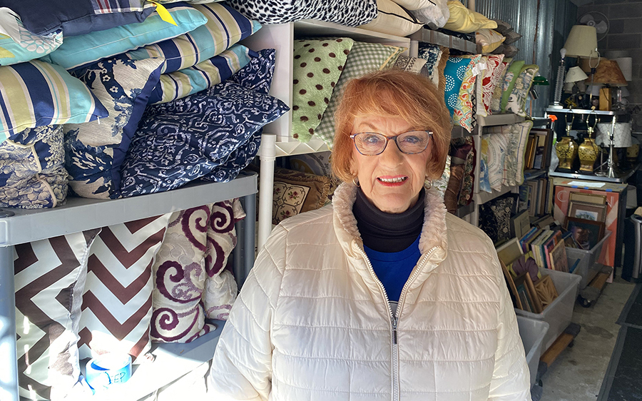 Gracing Spaces volunteer Roz Knapp stands in front of a shelving unit filled with decorative throw pillows.