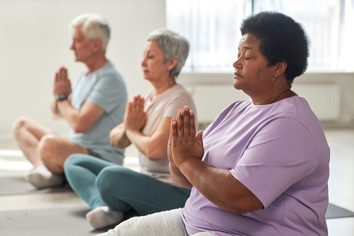 Three older adults sit on yoga mats with their legs crossed and their hands in prayer position.