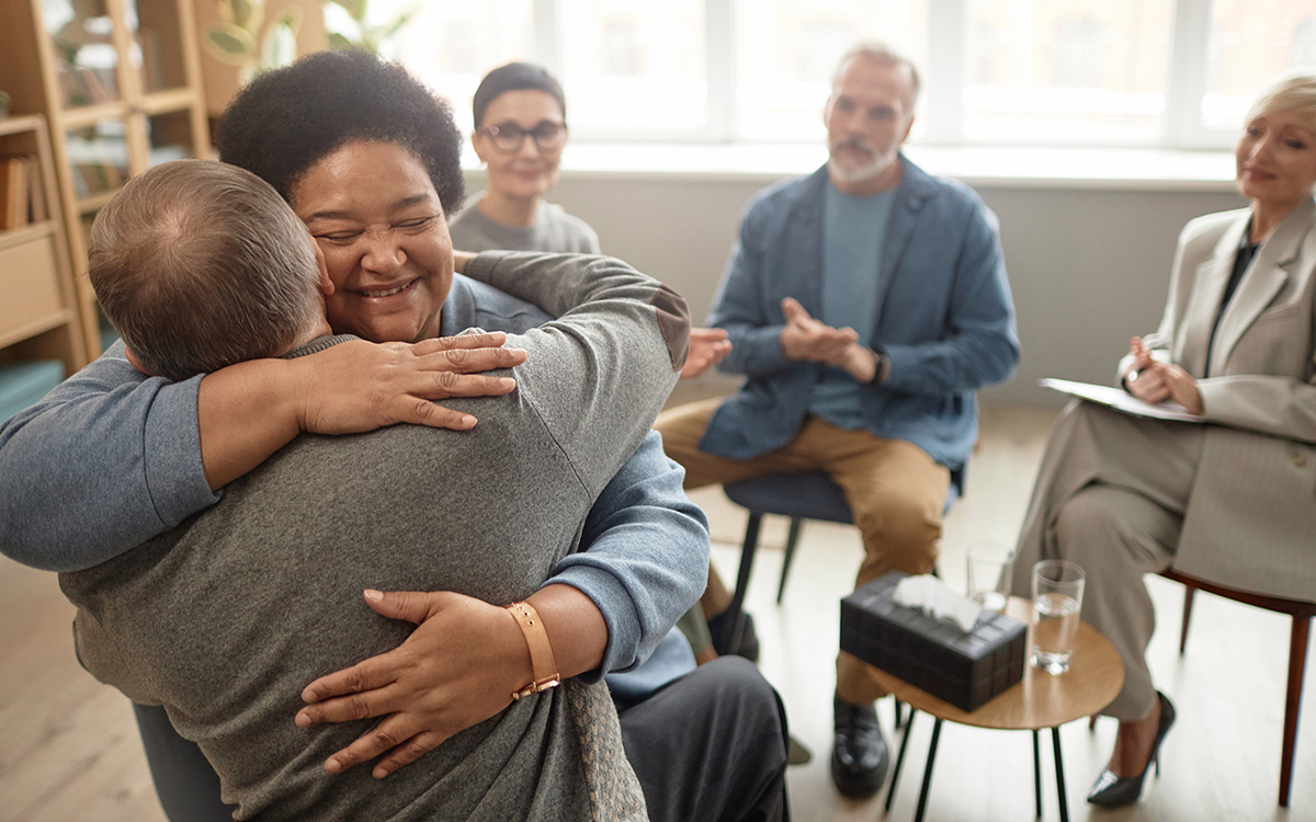 Two people hug while people in a group sit in the background.