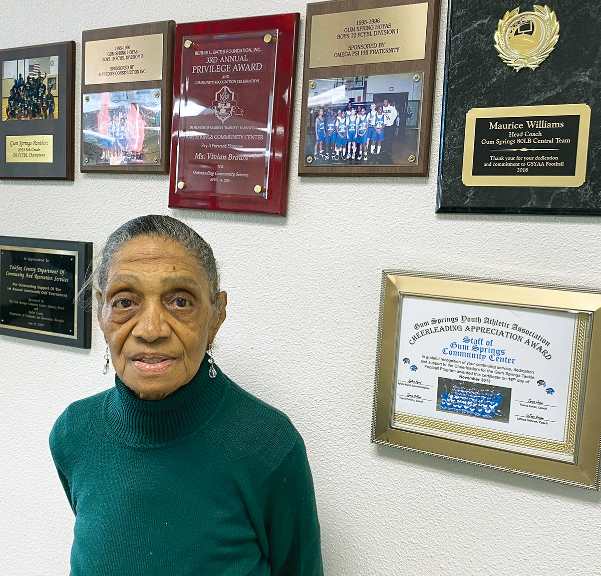 Vivian Brown standing in front of a wall at Gum Springs Community Center displaying various awards, including the President's Privilege Award she received for her volunteer work.