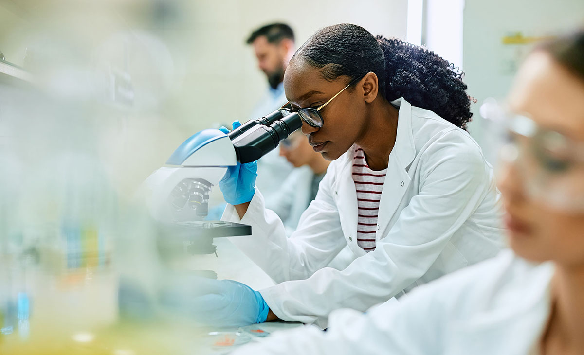 A young woman wearing a lab coat peers through a microscope while other researchers work nearby. 
