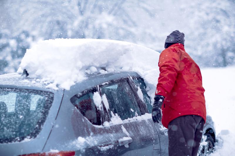Man in red jacket shovels snow off a parked vehicle while snow continues to fall