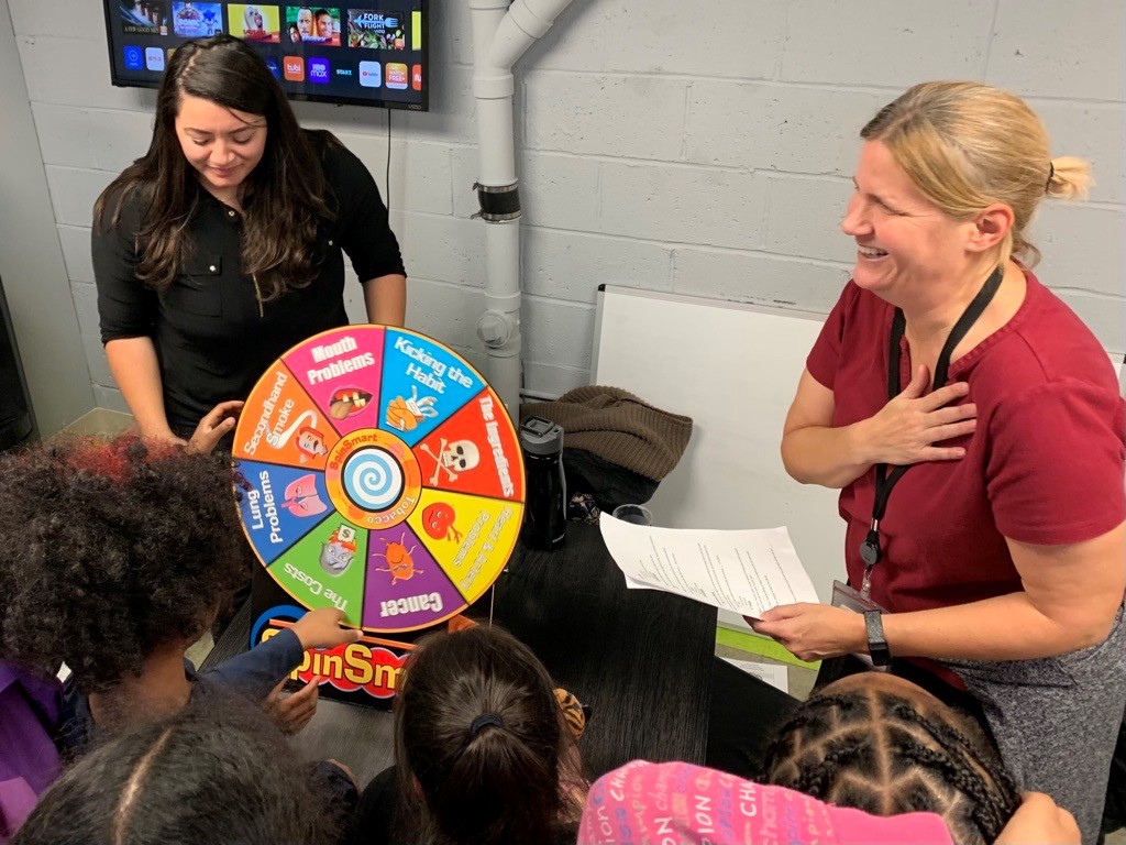 Two people showing children a colorful wheel with information on tobacco