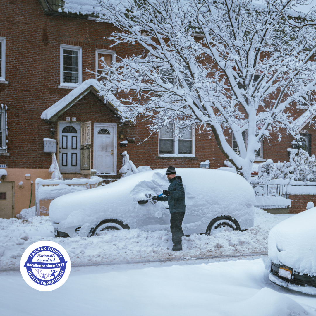 A man brushes snow off his snow-covered car on a neighborhood street. It is a cold winter day with heavy snowfall.