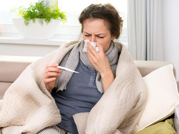 Woman sitting on sofa covering her nose and mouth with a tissue and reading a thermometer in her hand