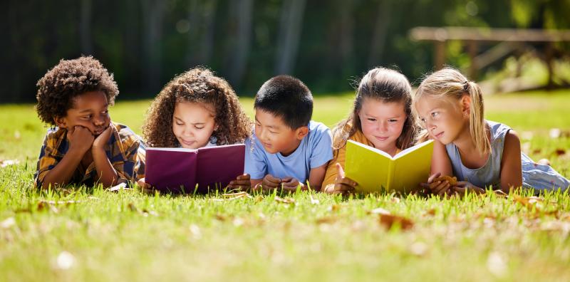 A group of 5 children read while laying on their stomachs in the grass