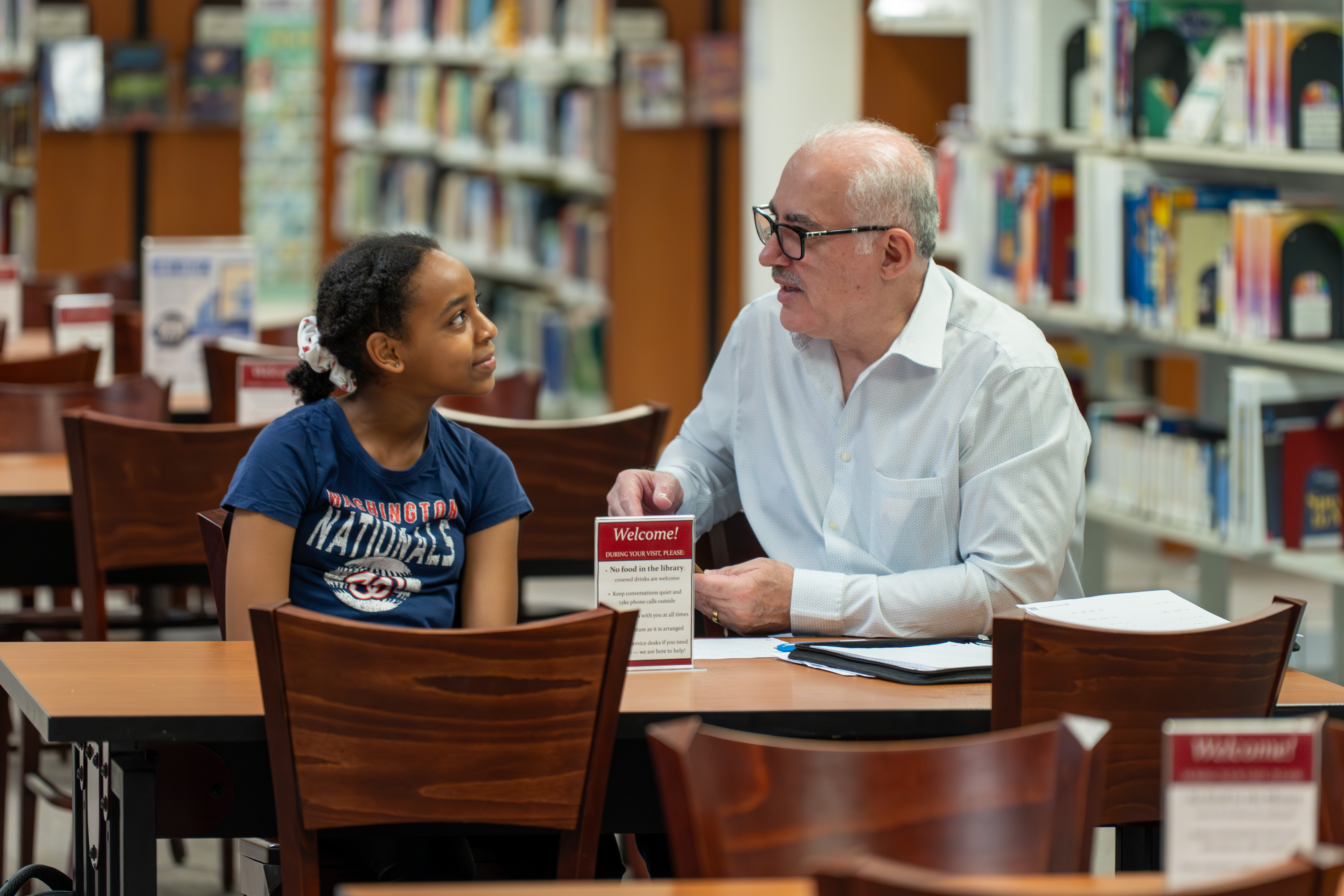 older man and young girl sit at a table discussing an assignment on a piece of paper