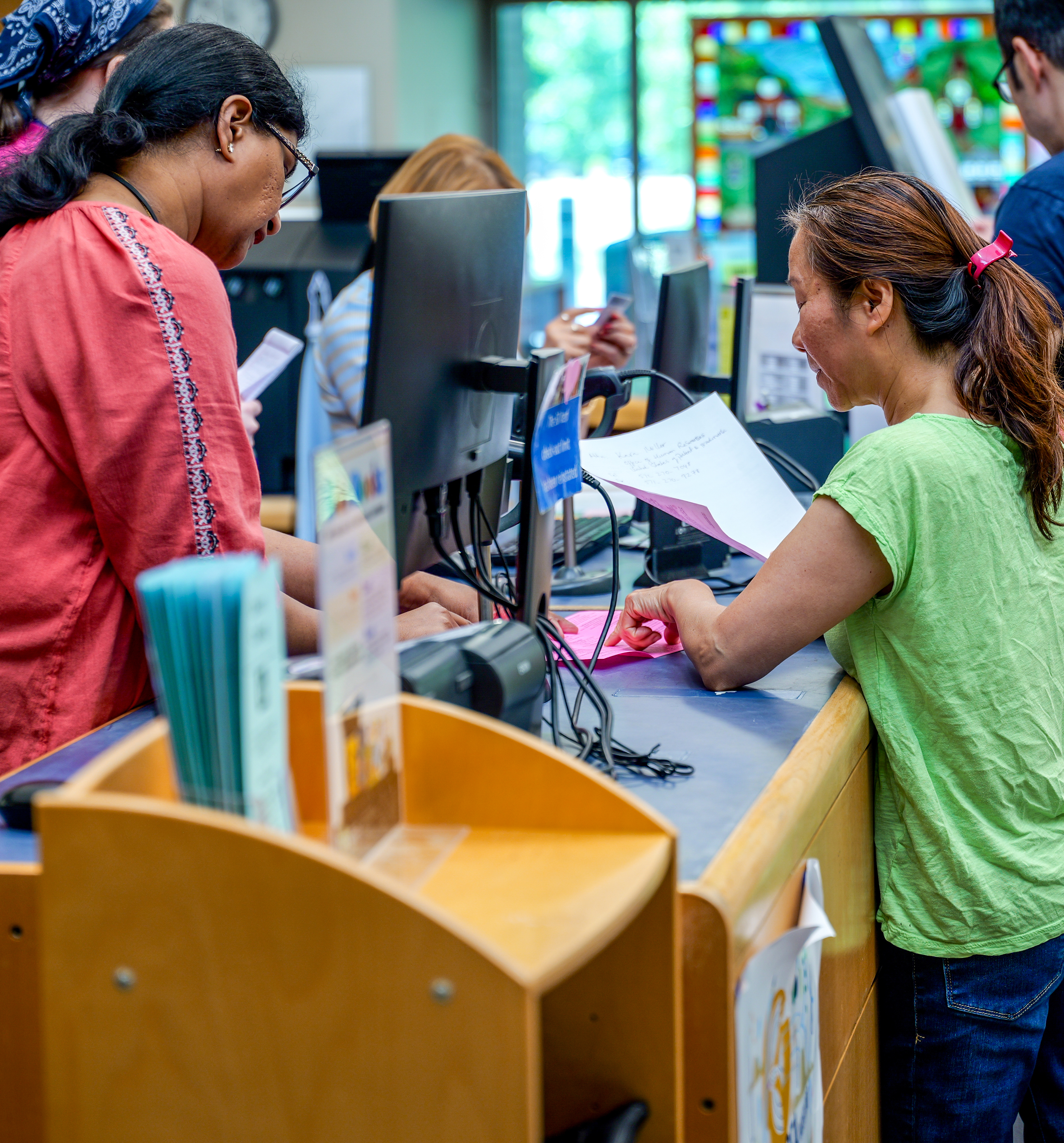 middle age asian woman talking to staff member behind the desk over a pink piece of paper