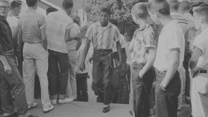 Black-and-white photo of a boy walking alone with books through a crowd of other students watching him.
