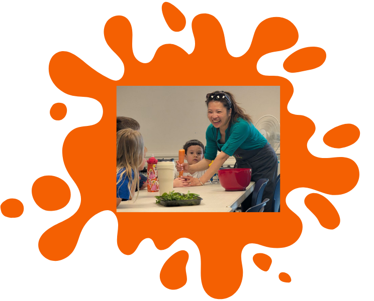 Middle aged Asian woman helps a young boy grate a carrot while smiling at other children. Framed in orange paint splat