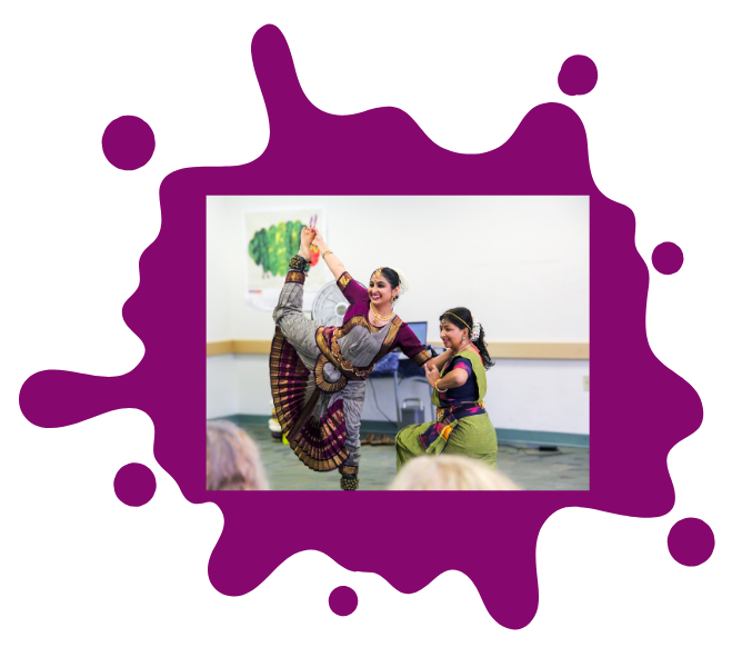 Middle aged Indian-descent woman, kneeling, in a green traditional outfit supports a young adult Indian-descent woman, wearing a purple traditional outfit, with leg lifted in dance move. Framed in a green paint splat