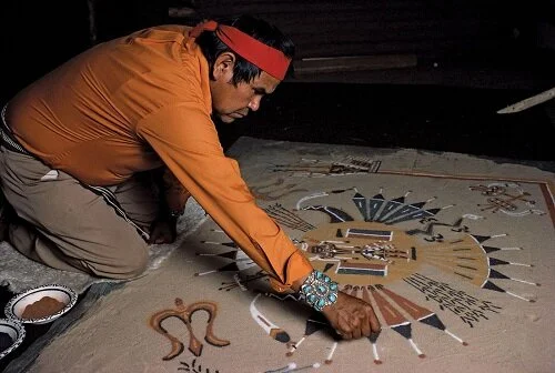 A man in traditional clothing and a red headband carefully creates an intricate sand painting on the ground.