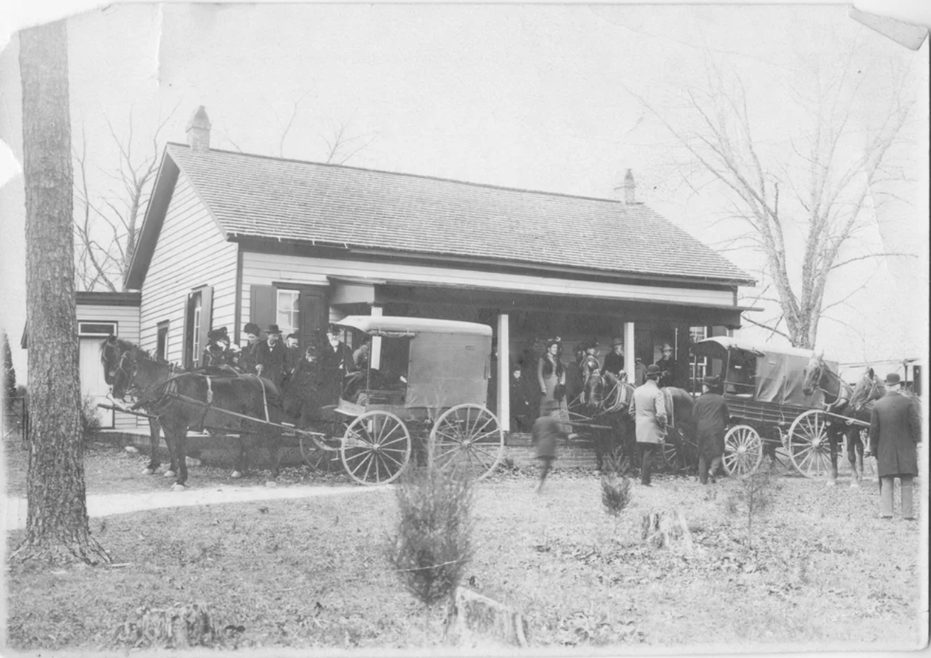 Black-and-white photo of people gathered outside a small house with horse-drawn carriages lined up in front.