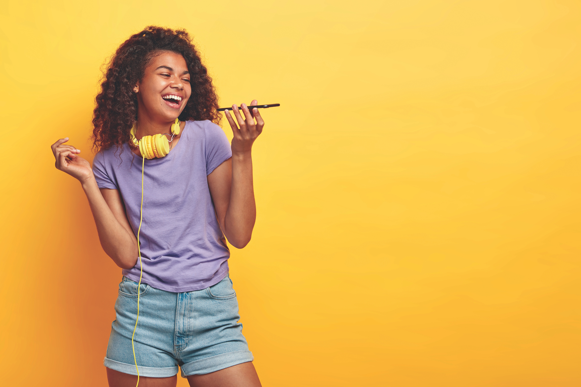 Smiling, Black teenage girl in a light purple t-shirt and jean shorts stands in front of a yellow background with headphones around her neck and holds a cell phone