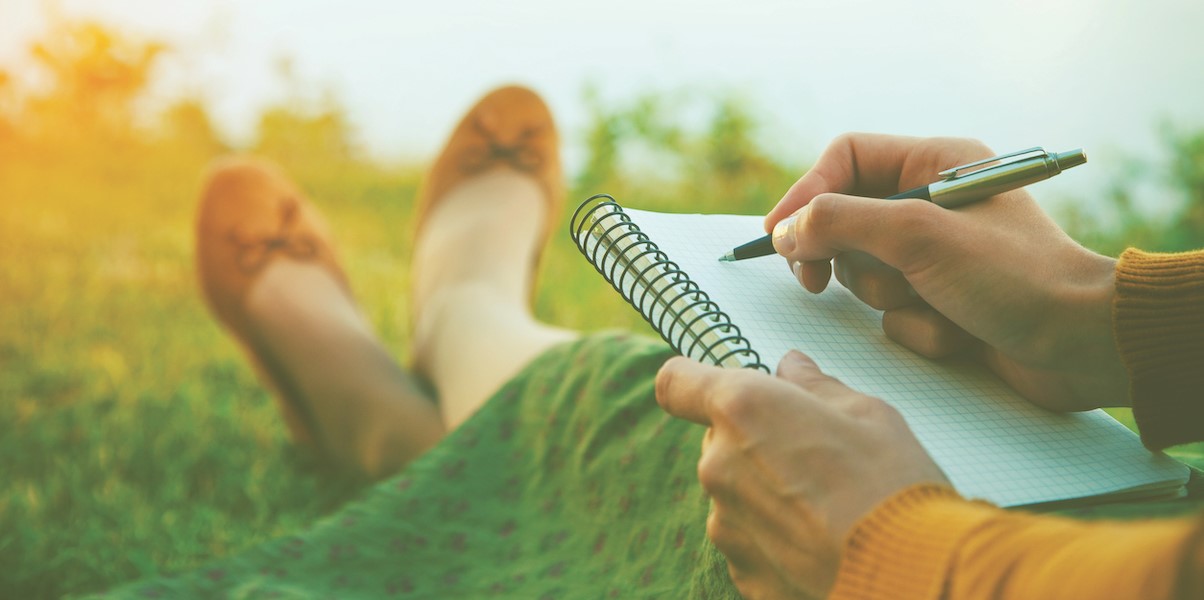 woman's hands hold a pen and journal in her lap while she sits in the grass