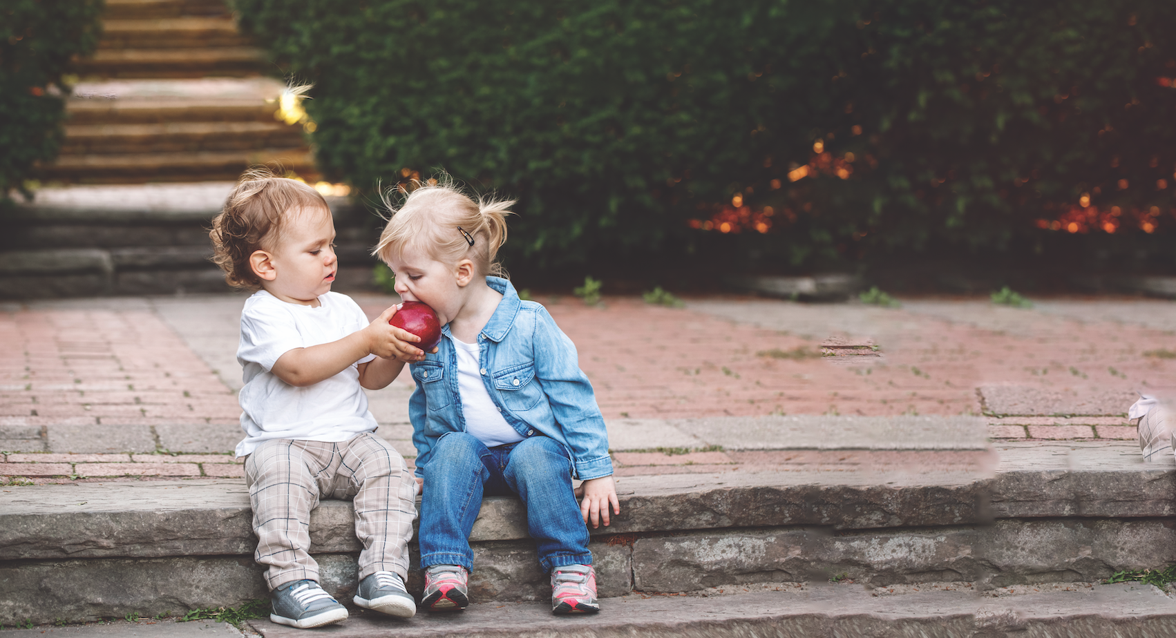 young boy and girl sit outside on a step and boy holds up apple while girl takes a bite