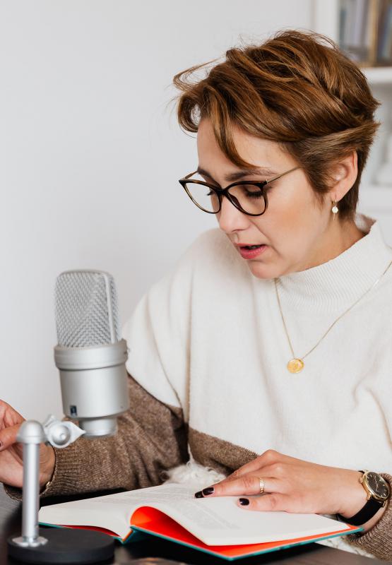 woman with sitting at a desk reading an open book into a microphone