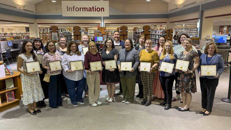A group of seven adults stands in a row inside a library in front of an “Information” sign, each person holding a framed certificate and facing the camera. Bookshelves and computer stations fill the background.