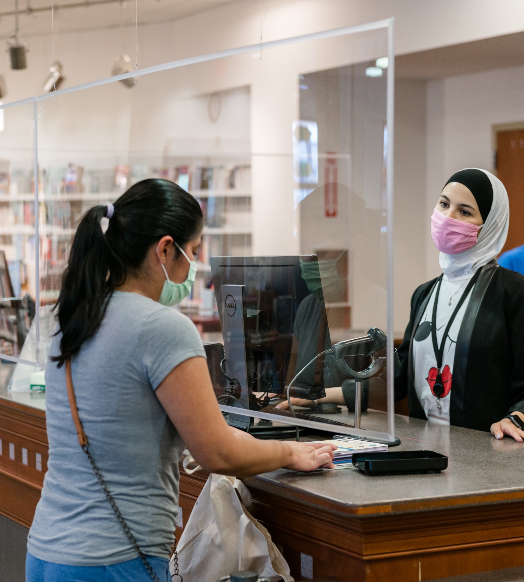 a girl in a gray shirt with black hair in a pony tail stands at the library info desk talking to a library staff member behind the desk 