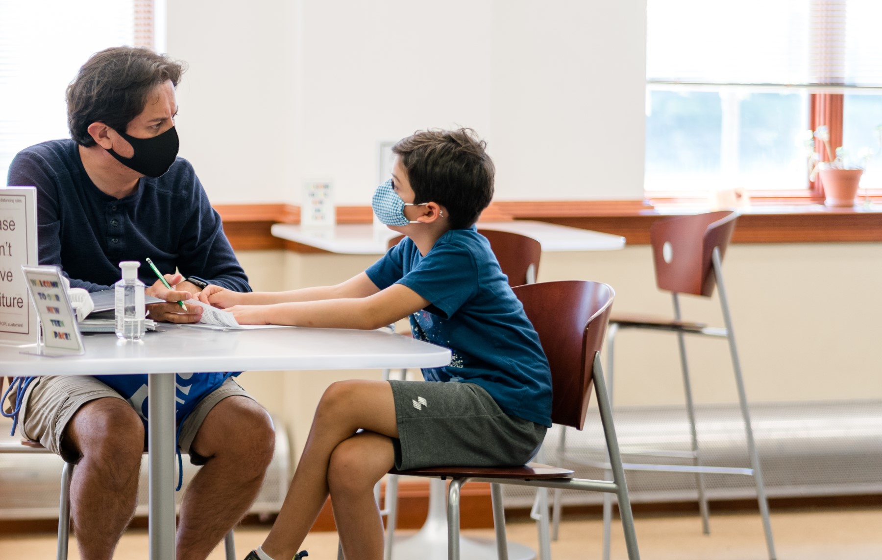 a man and boy both wearing masks sit at a table discussing an assignment on a piece of paper