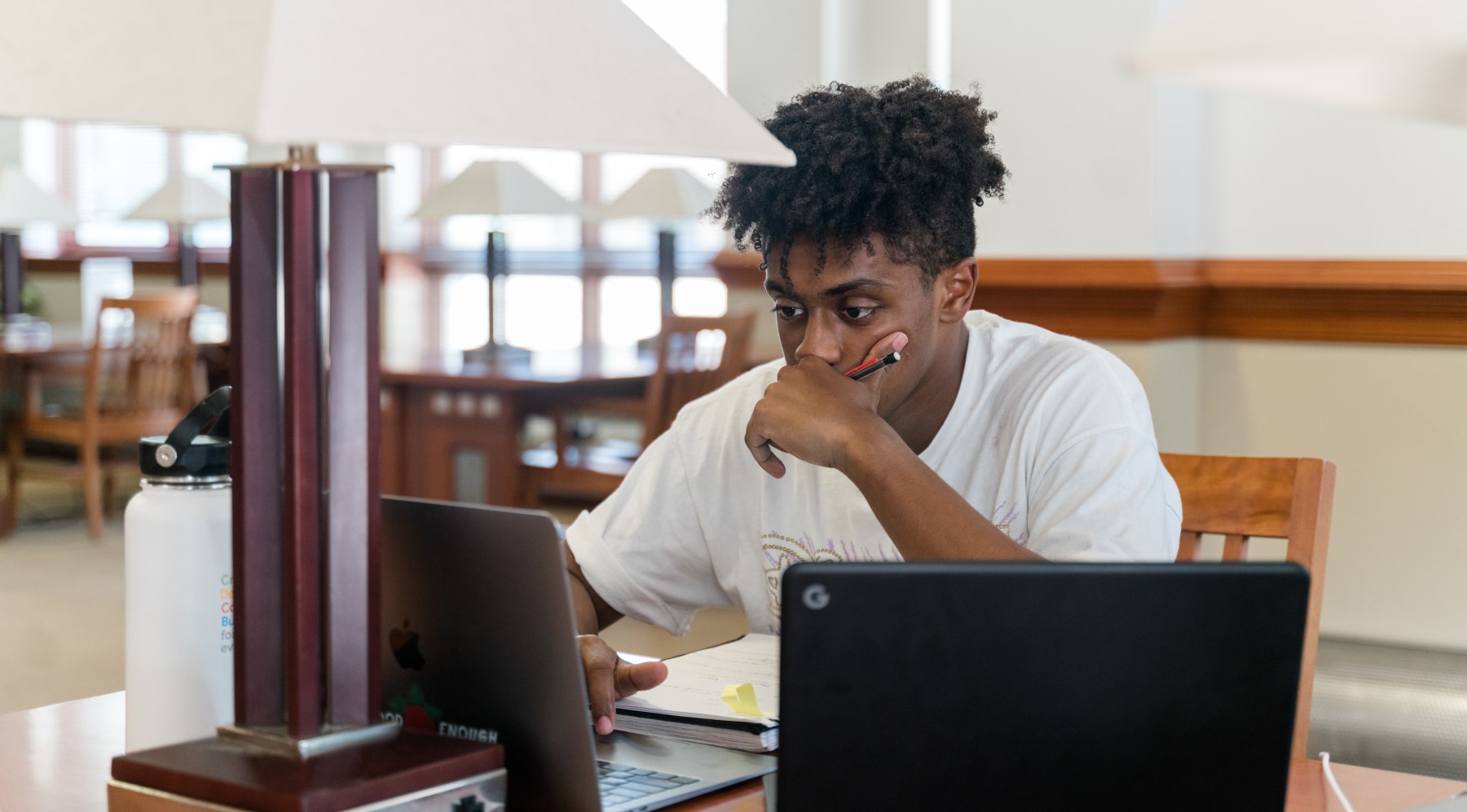 a young man sits at a library desk with two laptops open in front of him and a hand held pensively to his chin