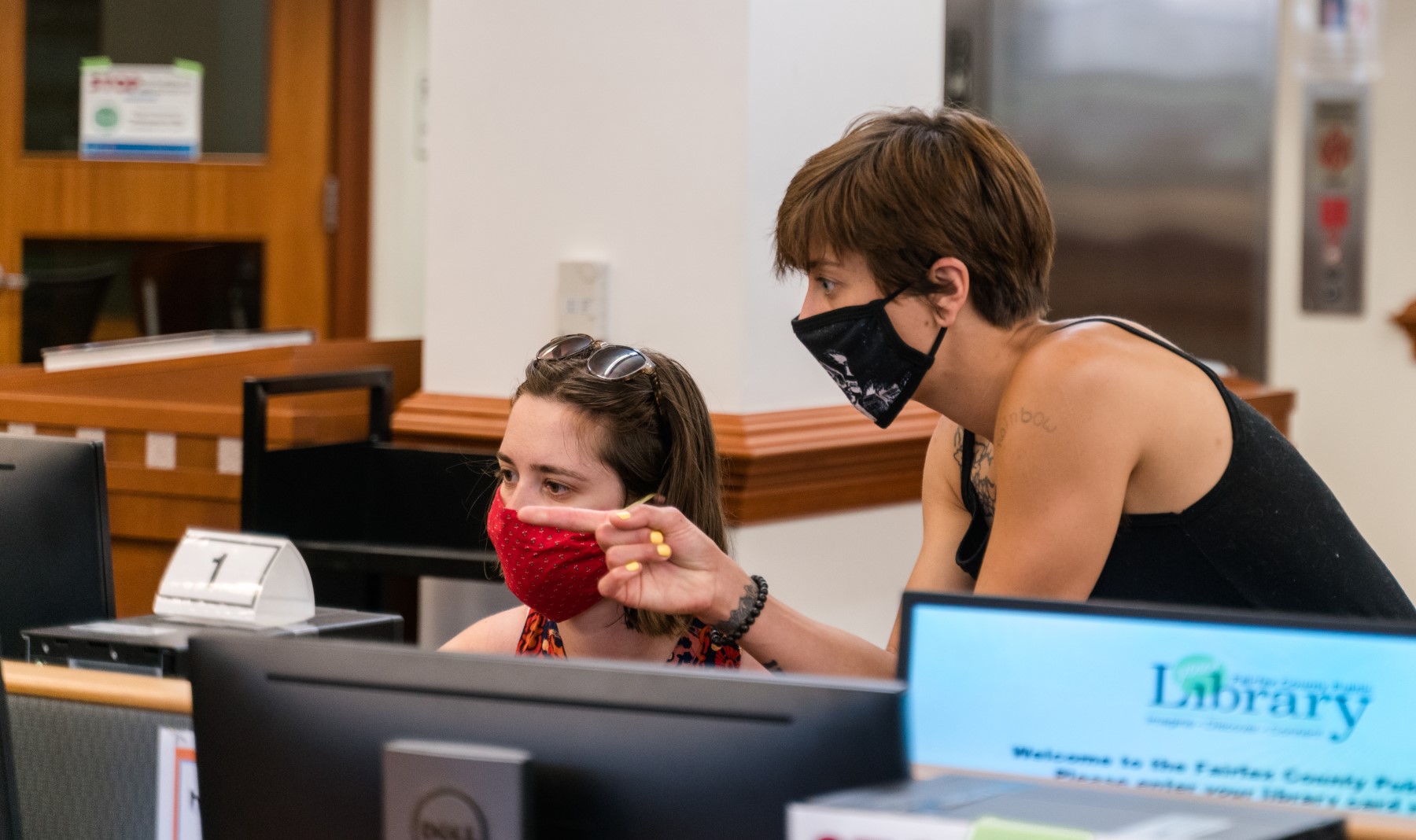 a young woman wearing a mask points over the shoulder of another young woman wearing a mask and sitting at a library desktop computer