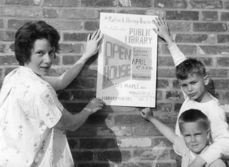 A woman and children holding a sign for Patrick Henry Library that states "Open House ... Everyone Welcome."