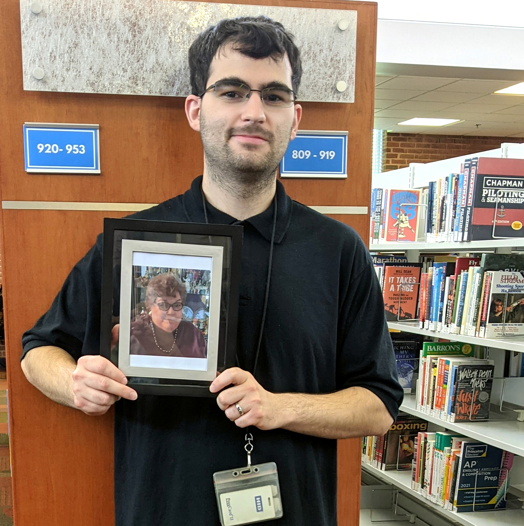 library intern Dylan with dark hair and glasses stands in front of library shelves of books and holds a photo of his grandmother