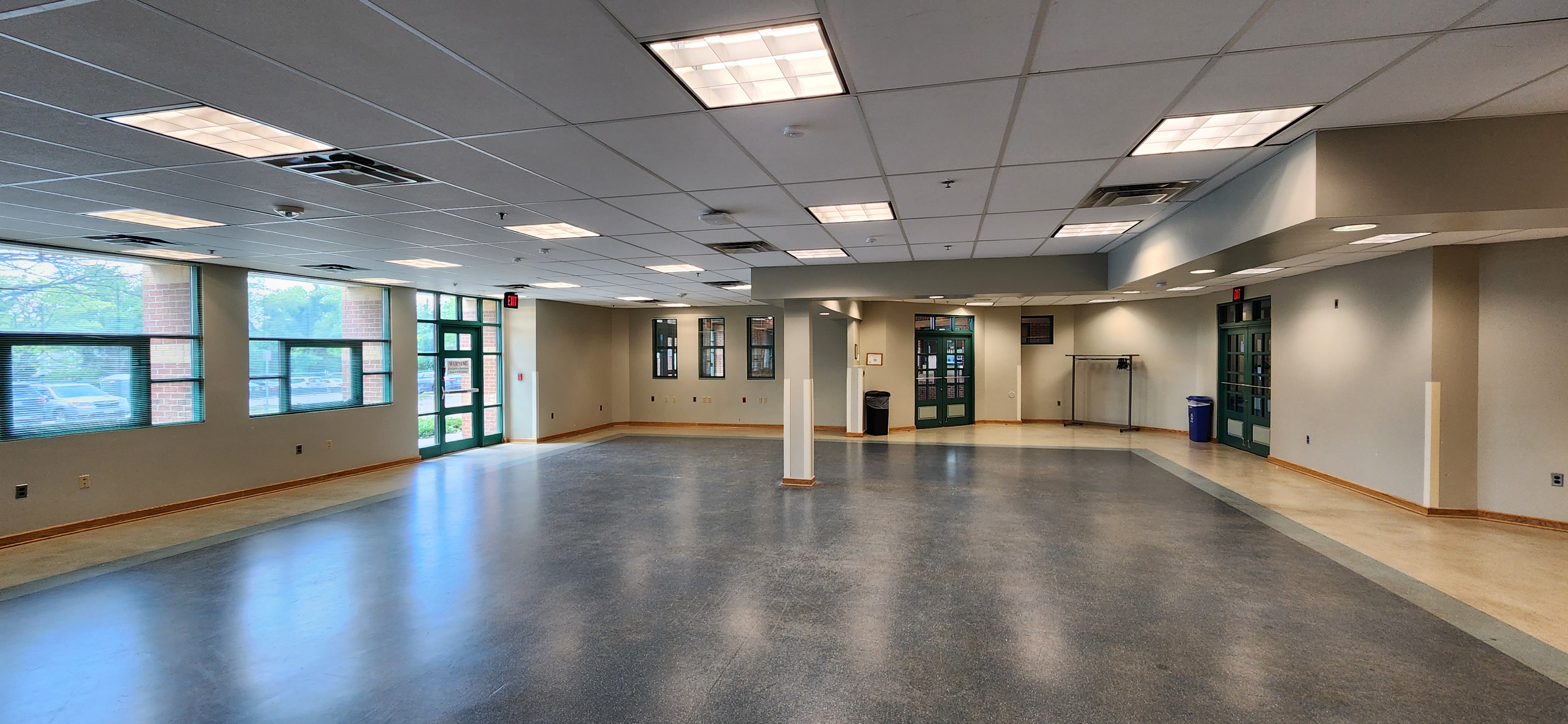 Open multipurpose room with large windows, central support columns, green double doors, and wide floor space for events or gatherings.