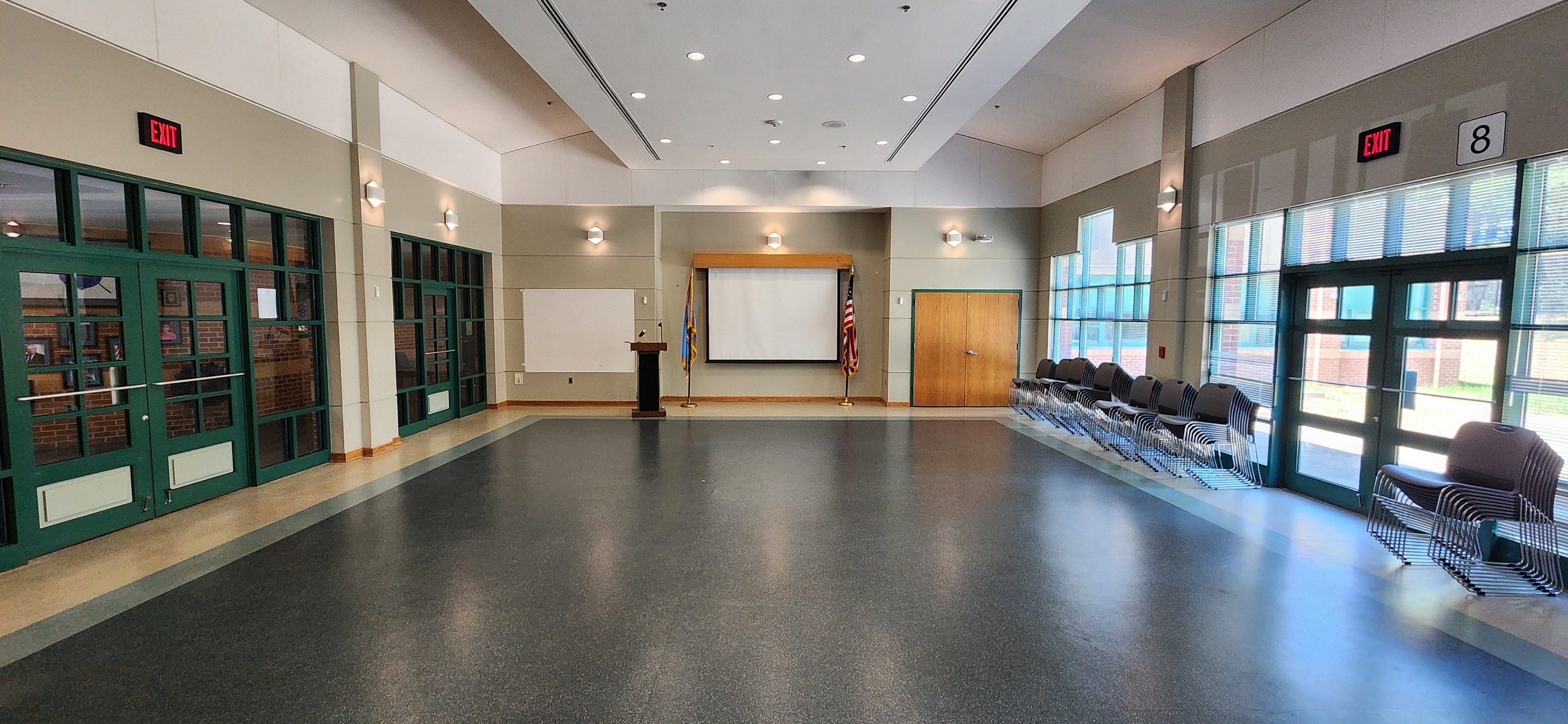 Wide, well-lit room with a podium and screen at the front, stacked chairs on one side, and tall windows and green doors around the space.